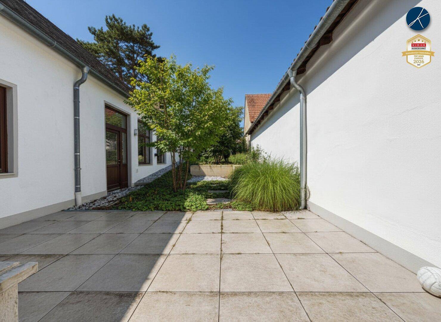 Sonniger Terrassenbereich zwischen zwei Gebäudeteilen mit Steinplatten und grüner Bepflanzung.