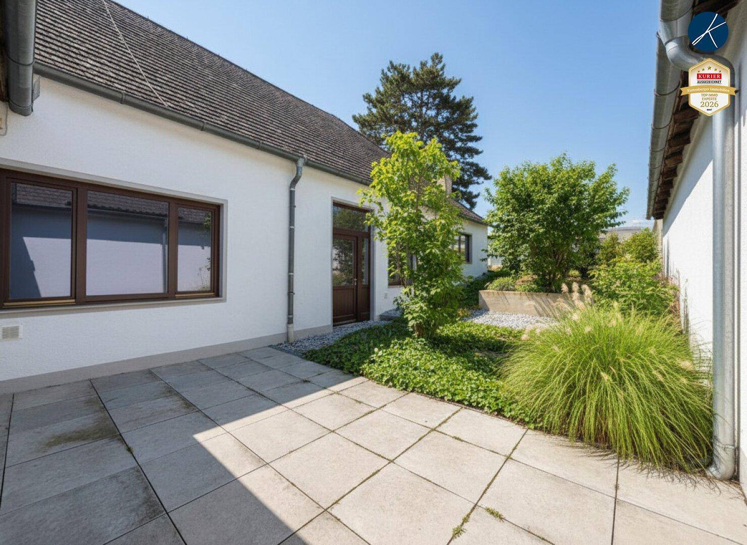 Sonniger Terrassenbereich mit Steinplatten und grüner Bepflanzung, angrenzend an das Gebäude.