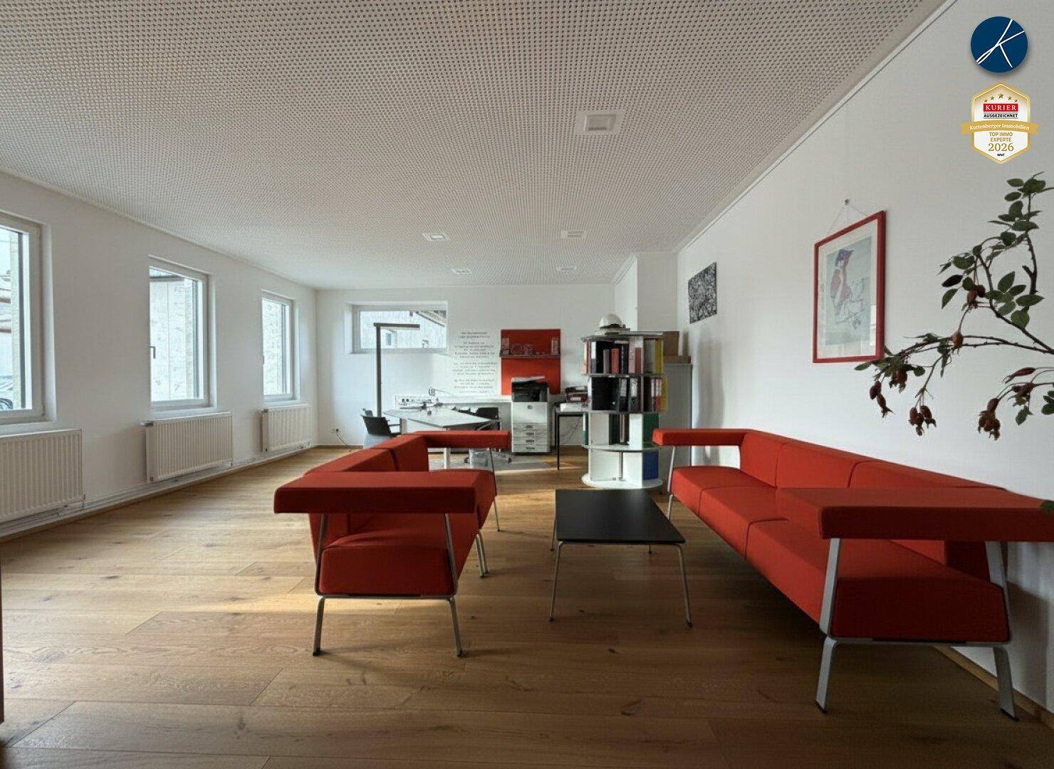 Heller Büroraum mit modernen roten Sofas, Holzboden und Blick auf den Arbeitsbereich im Hintergrund.