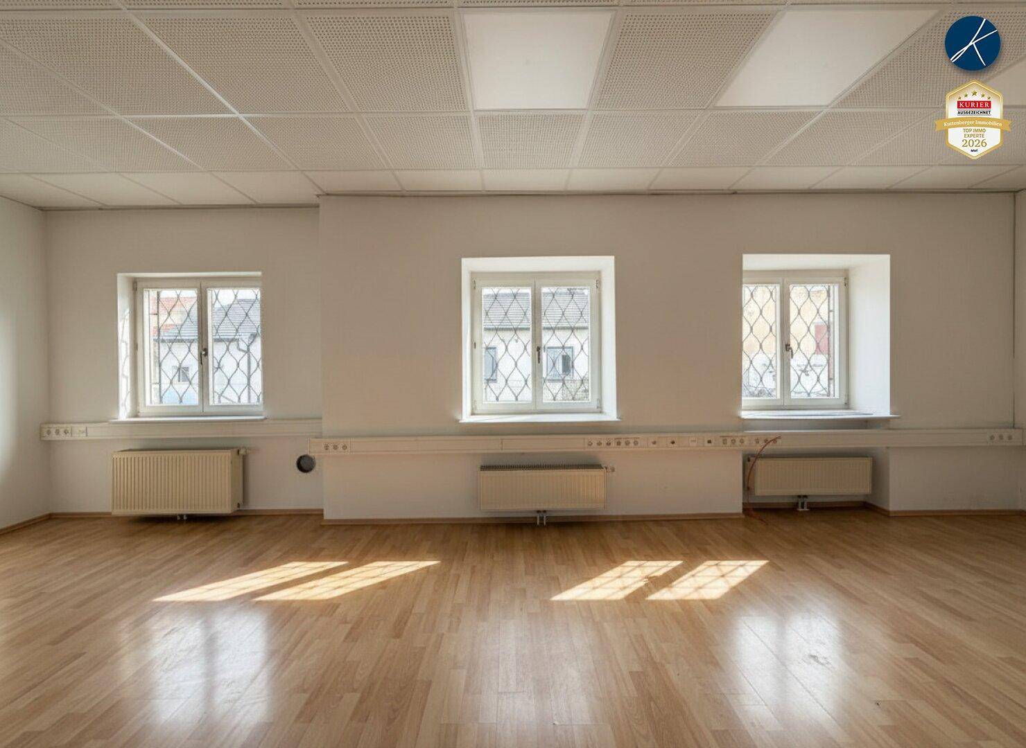 Leerer Raum mit Holzboden, drei Fenstern und Heizkörpern unter der Fensterbank, gut beleuchtet.