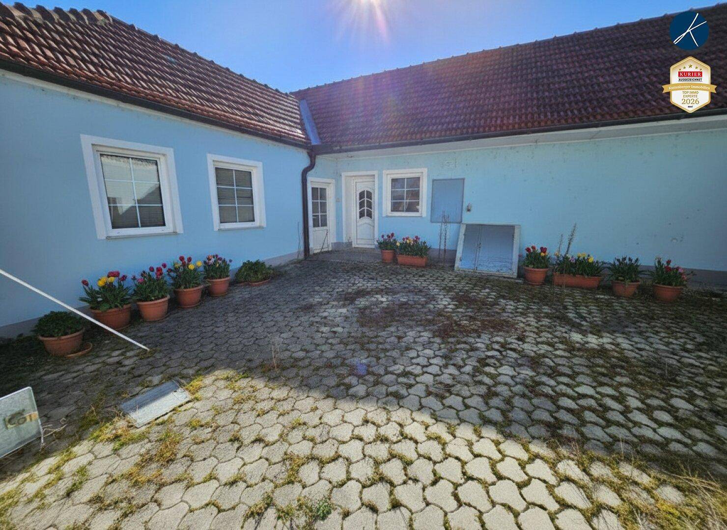 Gepflasterter Innenhofbereich neben dem Haus, geschmückt mit bunten Topfblumen in der Sonne.