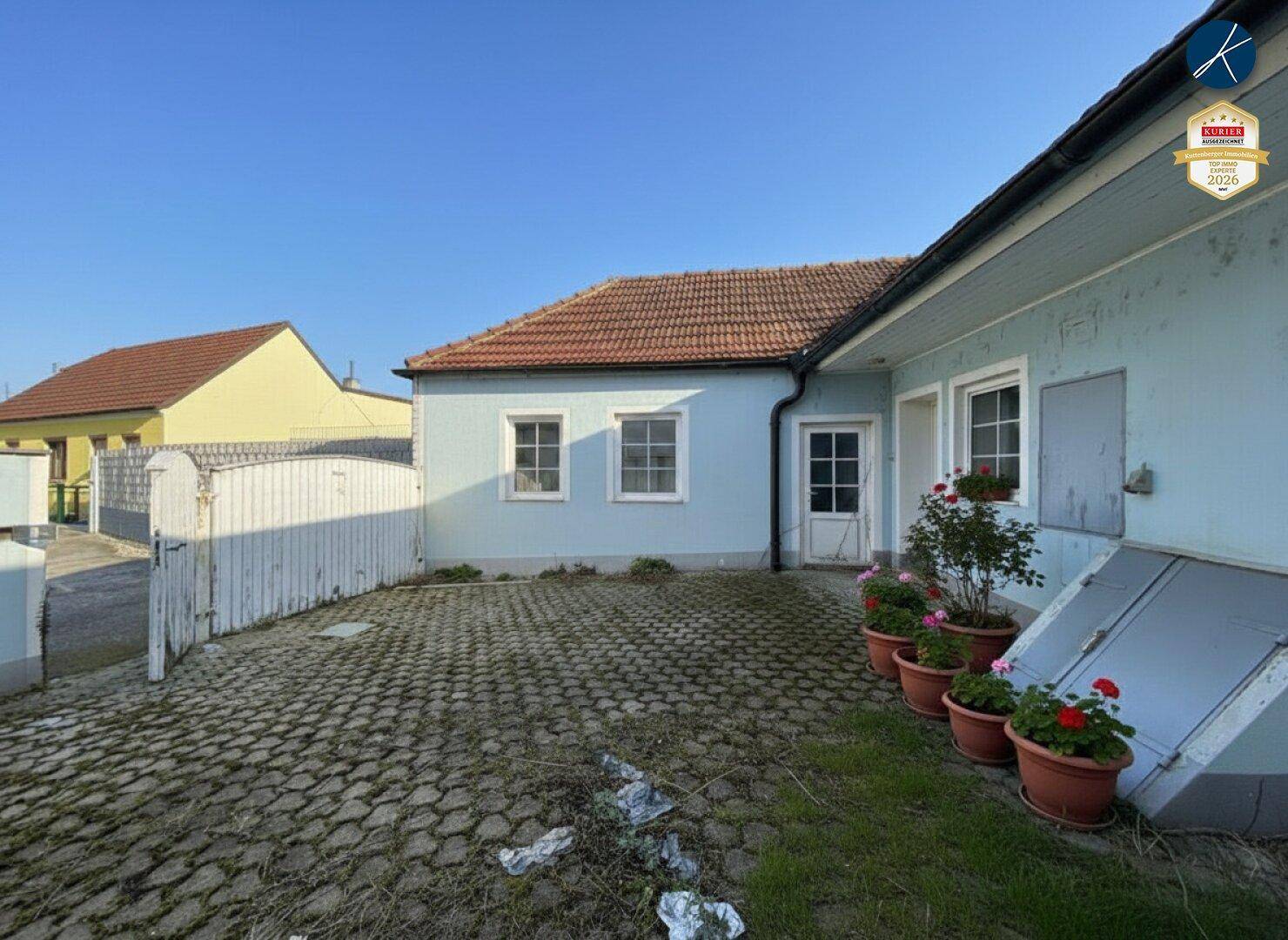 Sonniger gepflasterter Hofbereich mit Topfblumen und einem weißen Zaun, der den Eingang begrenzt.