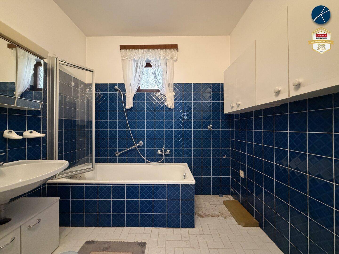 Badezimmer mit blauen Fliesen, Badewanne mit Duschabtrennung und Fenster in einem 80er-Jahre-Stil.