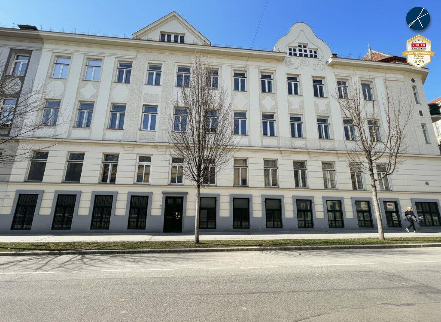 Breite Fassade eines mehrstöckigen Gebäudes mit vielen Fenstern und Bäumen davor.