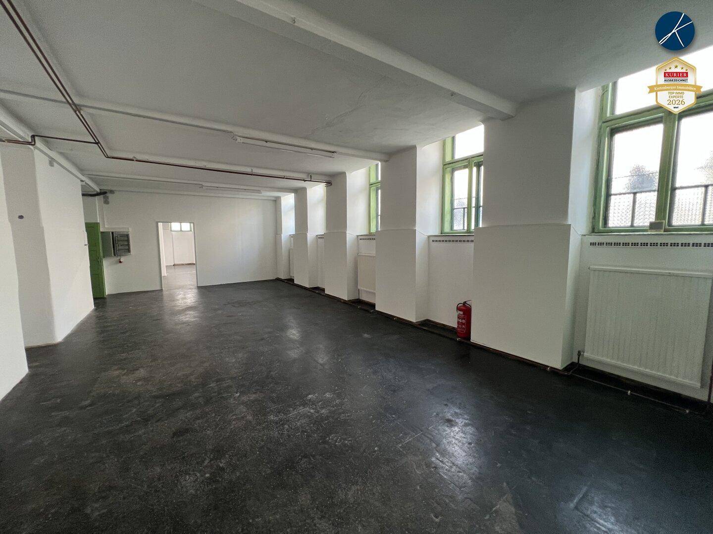 Weitläufiger Gewerberaum mit dunklem Boden, weißen Wänden und mehreren Fenstern für Tageslicht.