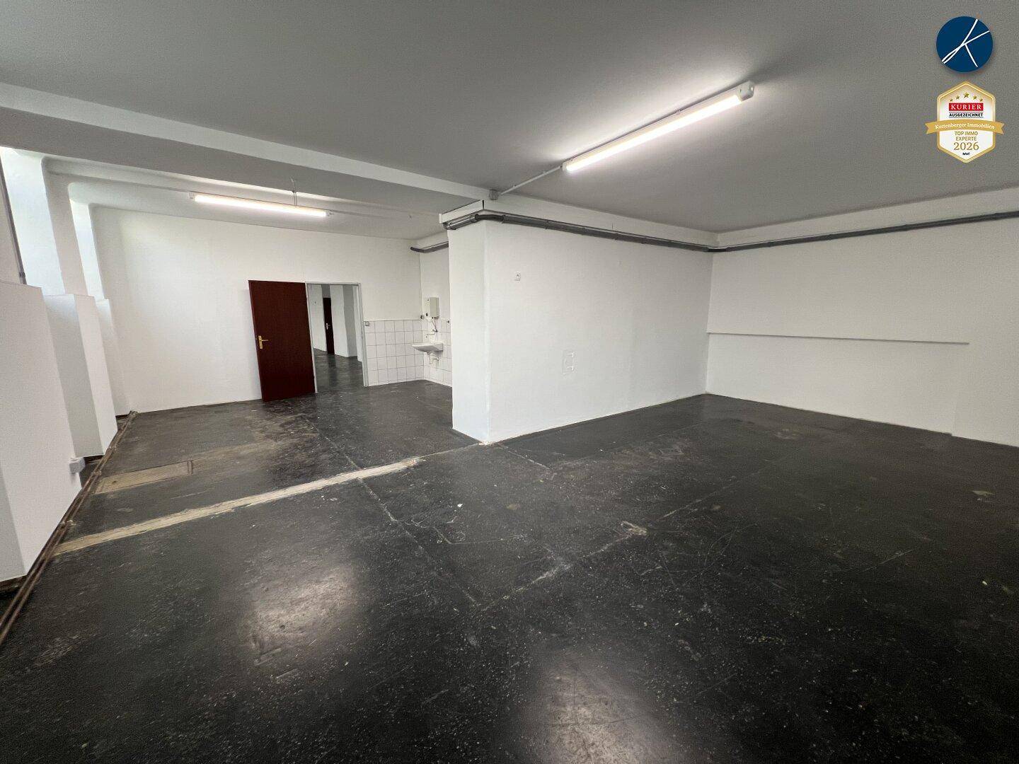 Großer, leerer Gewerberaum mit Waschbecken in der Ecke und dunklem Industrieboden.