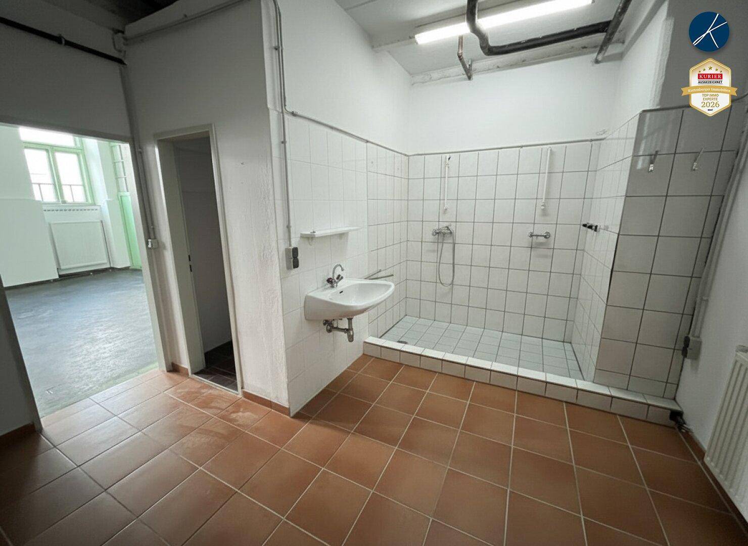 Funktionaler Duschraum mit mehreren Duschköpfen, Waschbecken und weißen Fliesen an den Wänden.