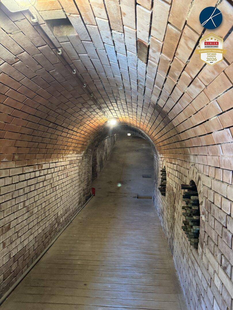 Langer, gewölbter Kellergang mit Ziegelwänden und Holzfußboden, beleuchtet von Deckenlampen.