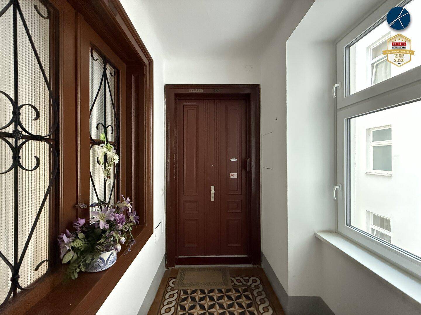Eingangstür einer Wohnung in einem Flur mit Fenster und dekorativem Gitter, das Tageslicht spendet.