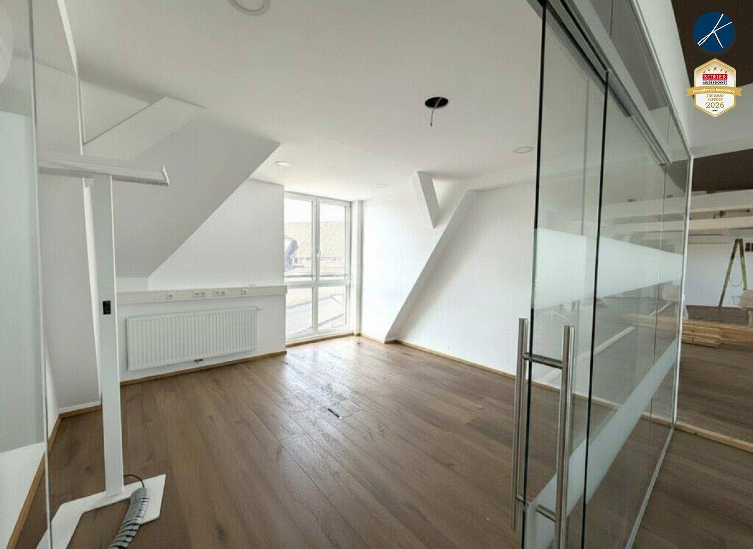Heller Büroraum mit Glasabtrennung, großem Fenster und hochwertigem Holzboden im Dachgeschoss.