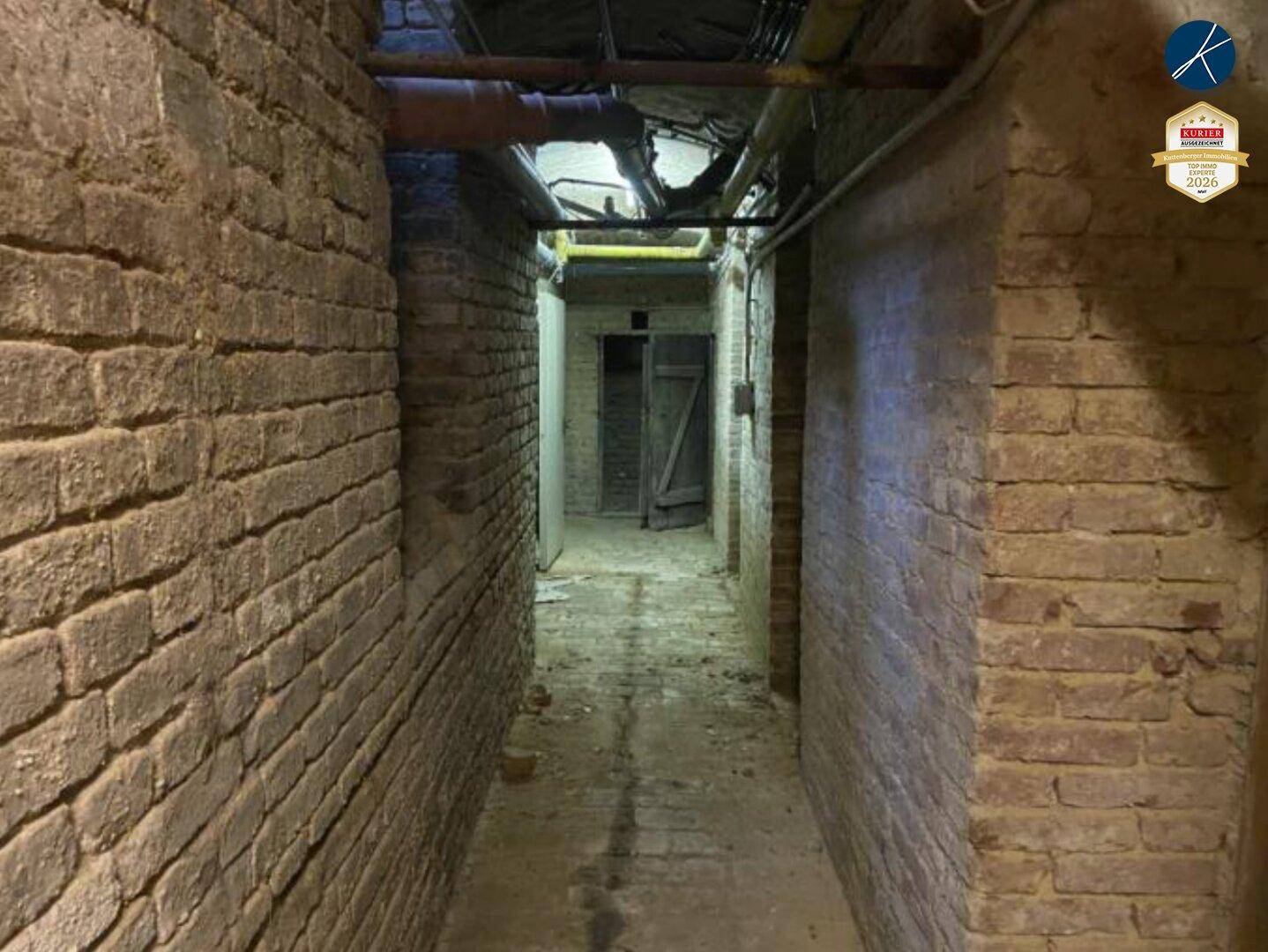 Langer, schmaler Kellergang mit unverputzten Ziegelwänden und sichtbaren Rohren an der Decke.