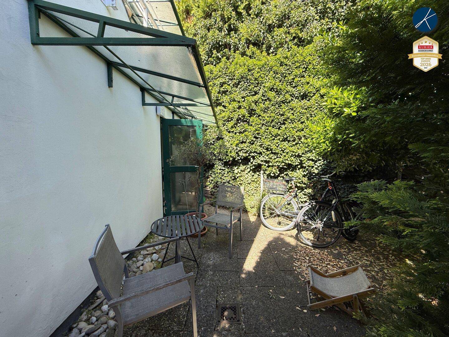 Kleine gepflasterte Terrasse mit Gartenmöbeln und üppiger grüner Bepflanzung im Hintergrund.