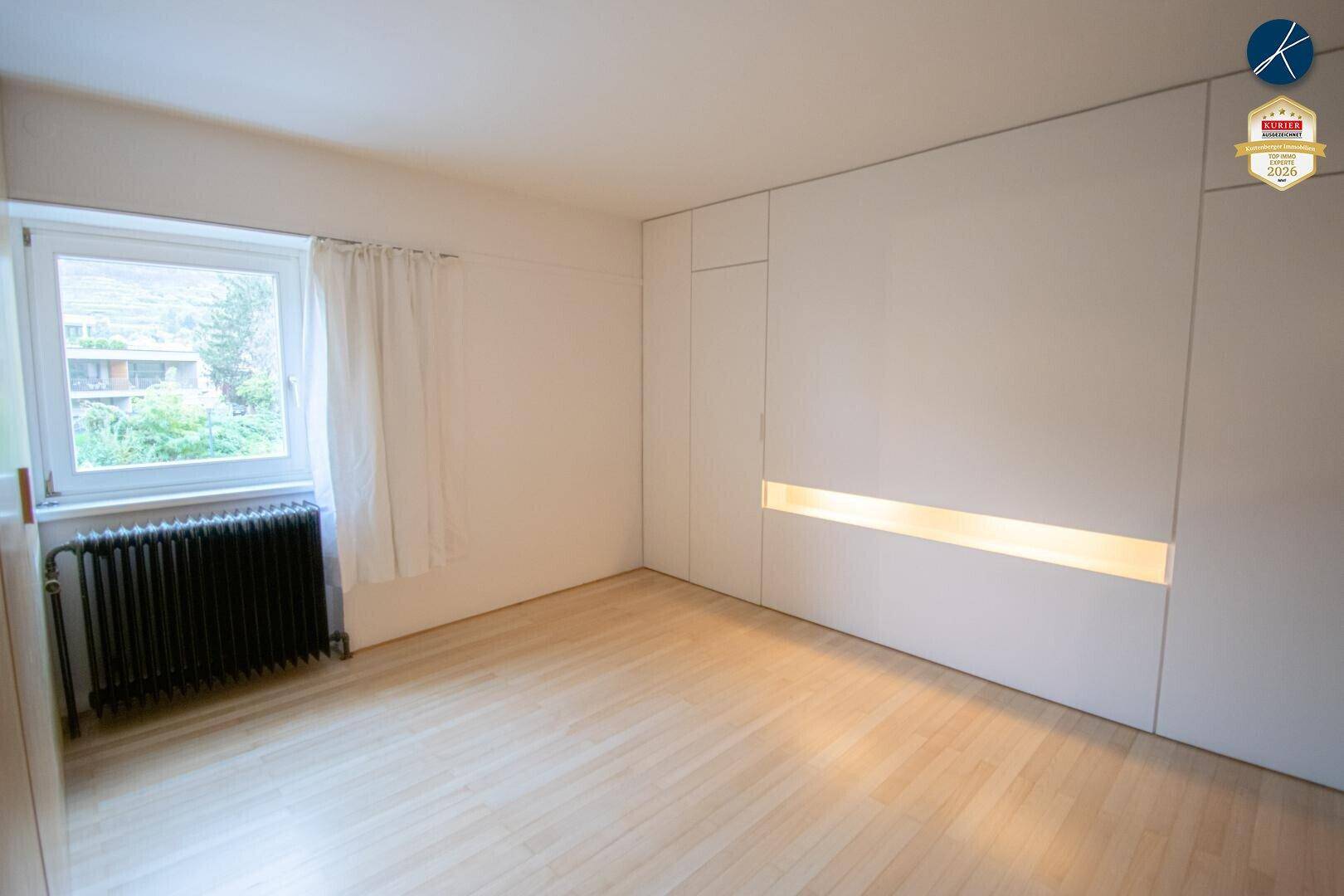 Helles Schlafzimmer mit Holzboden, großem Fenster und einer modernen Wand mit integrierter Beleuchtung.