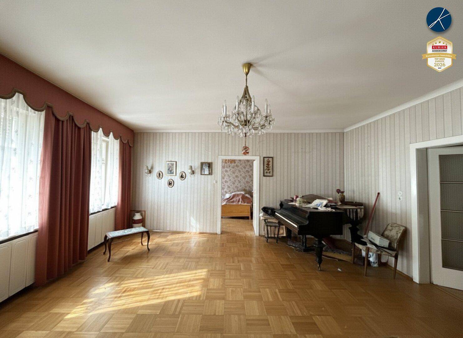 Geräumiges Wohnzimmer mit Fischgrätparkett, einem Flügel und traditioneller Einrichtung, mit Zugang zu einem weiteren Raum.