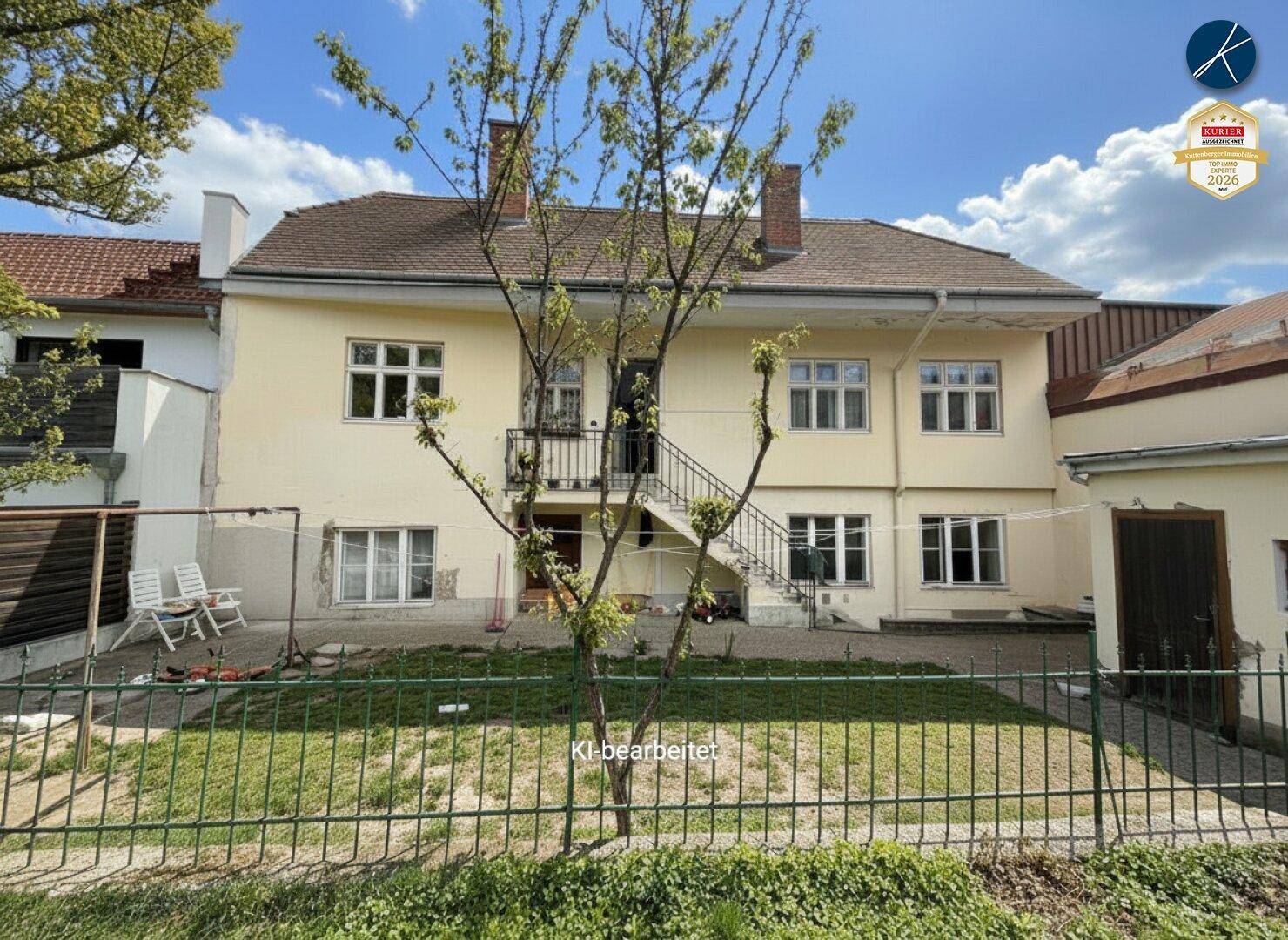 Gelbes Altbaugebäude mit Außentreppe und einem kleinen Gartenbereich im Vordergrund unter blauem Himmel.