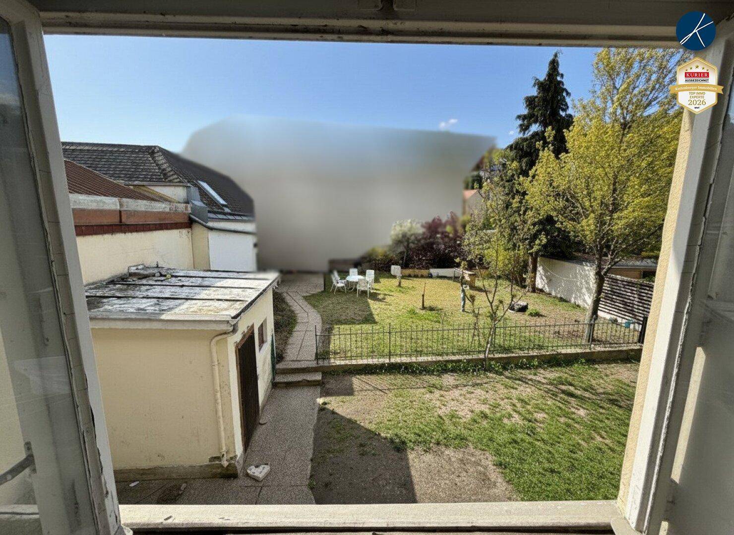 Blick aus einem Fenster auf einen Hinterhofgarten mit Rasenfläche, einem Schuppen und Gartenmöbeln.