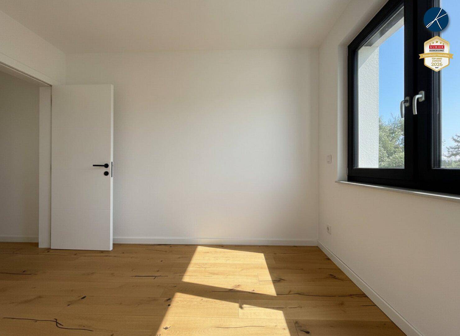 Helles, leeres Schlafzimmer mit Holzboden und großem Fenster, das viel Licht hereinlässt.
