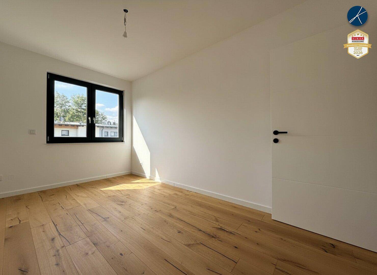 Geräumiges, leeres Zimmer mit Holzboden und Fenster, das von der Sonne durchflutet wird.