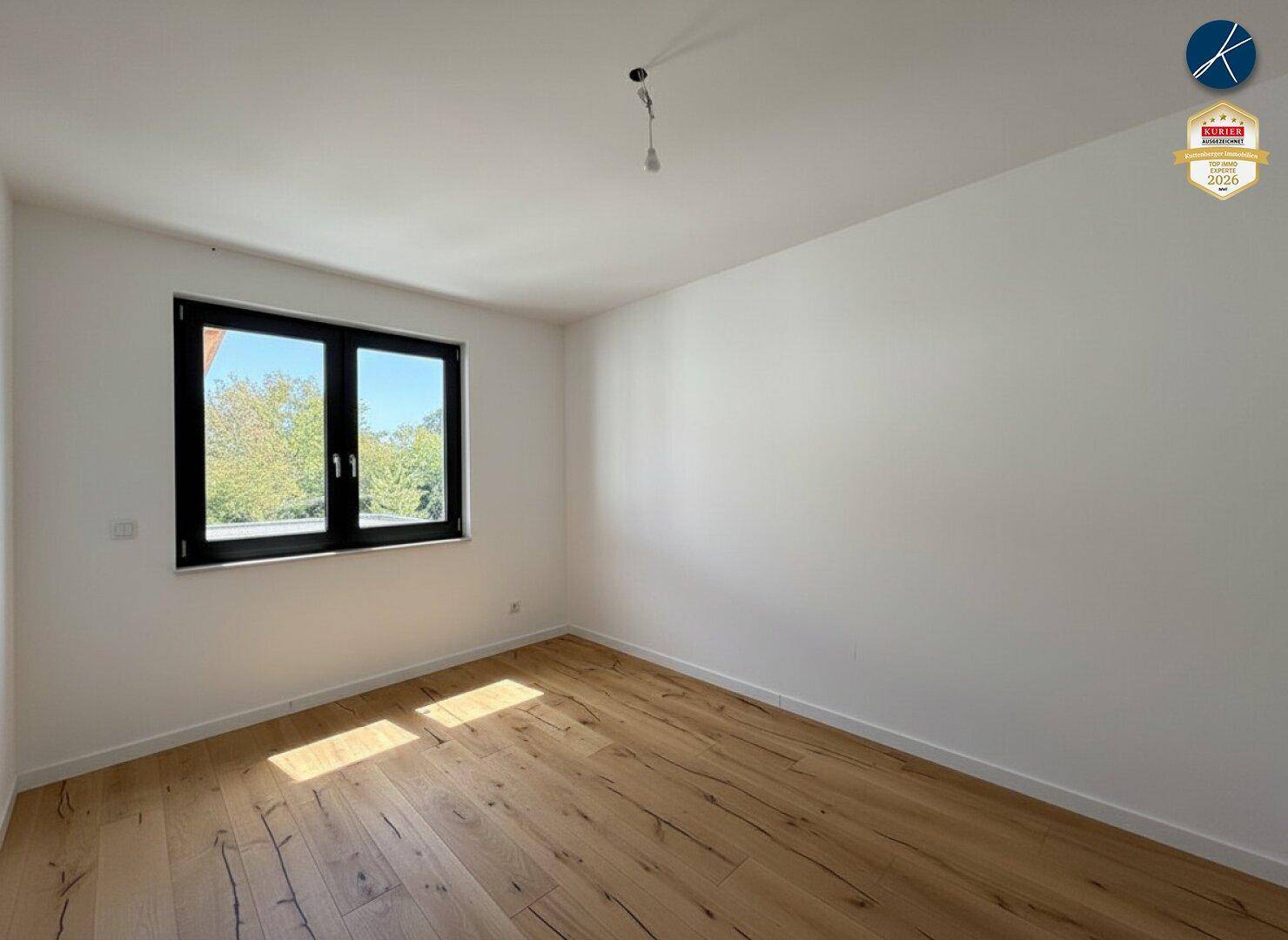 Schlichtes, leeres Zimmer mit Holzboden und einem Fenster, das einen Blick nach draußen bietet.