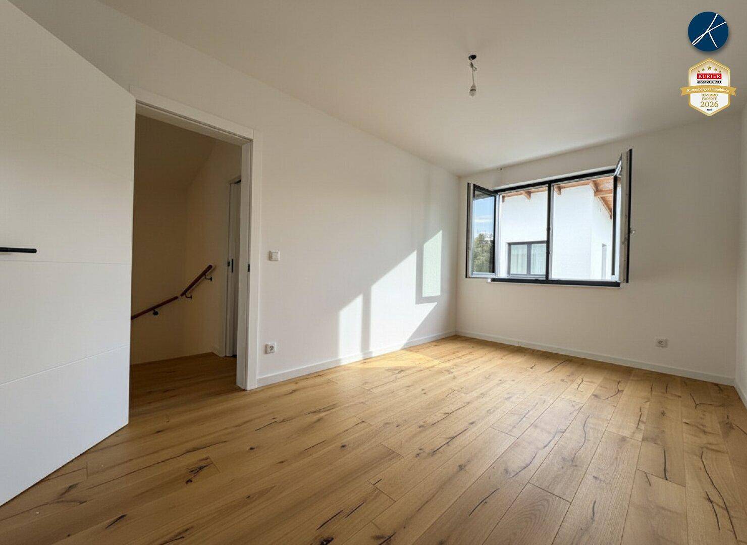 Helles, leeres Zimmer mit Holzboden und offenem Fenster, das frische Luft hereinlässt.