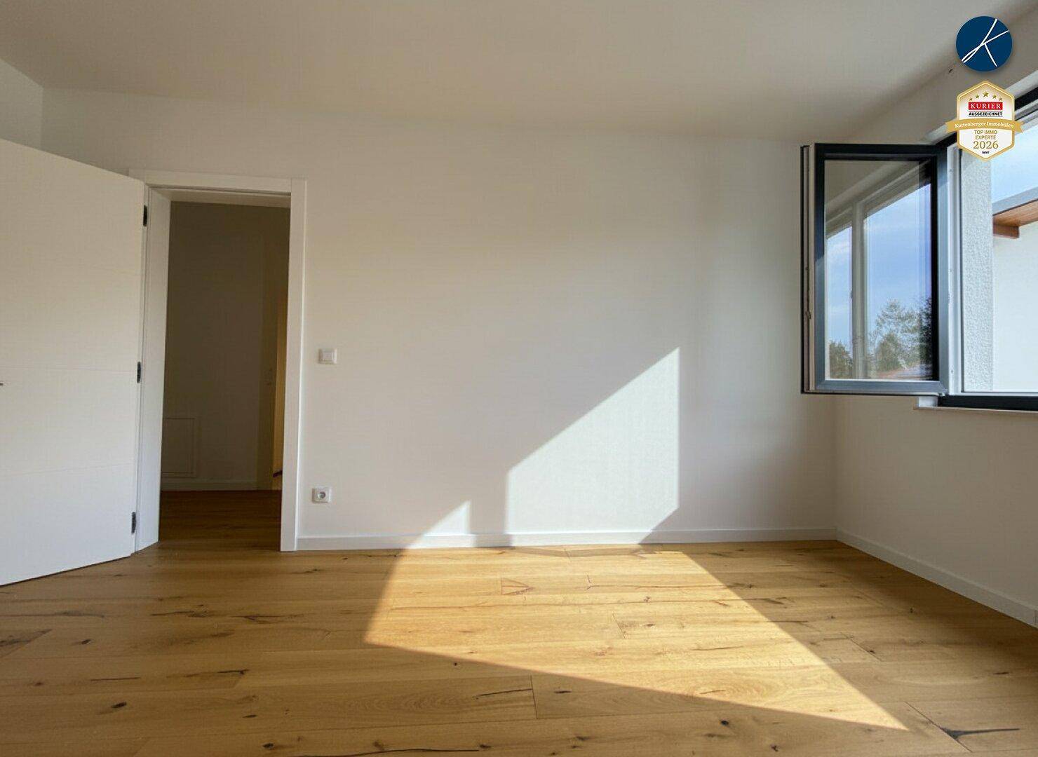 Geräumiges, leeres Zimmer mit Holzboden und geöffnetem Fenster, das viel Sonnenlicht empfängt.