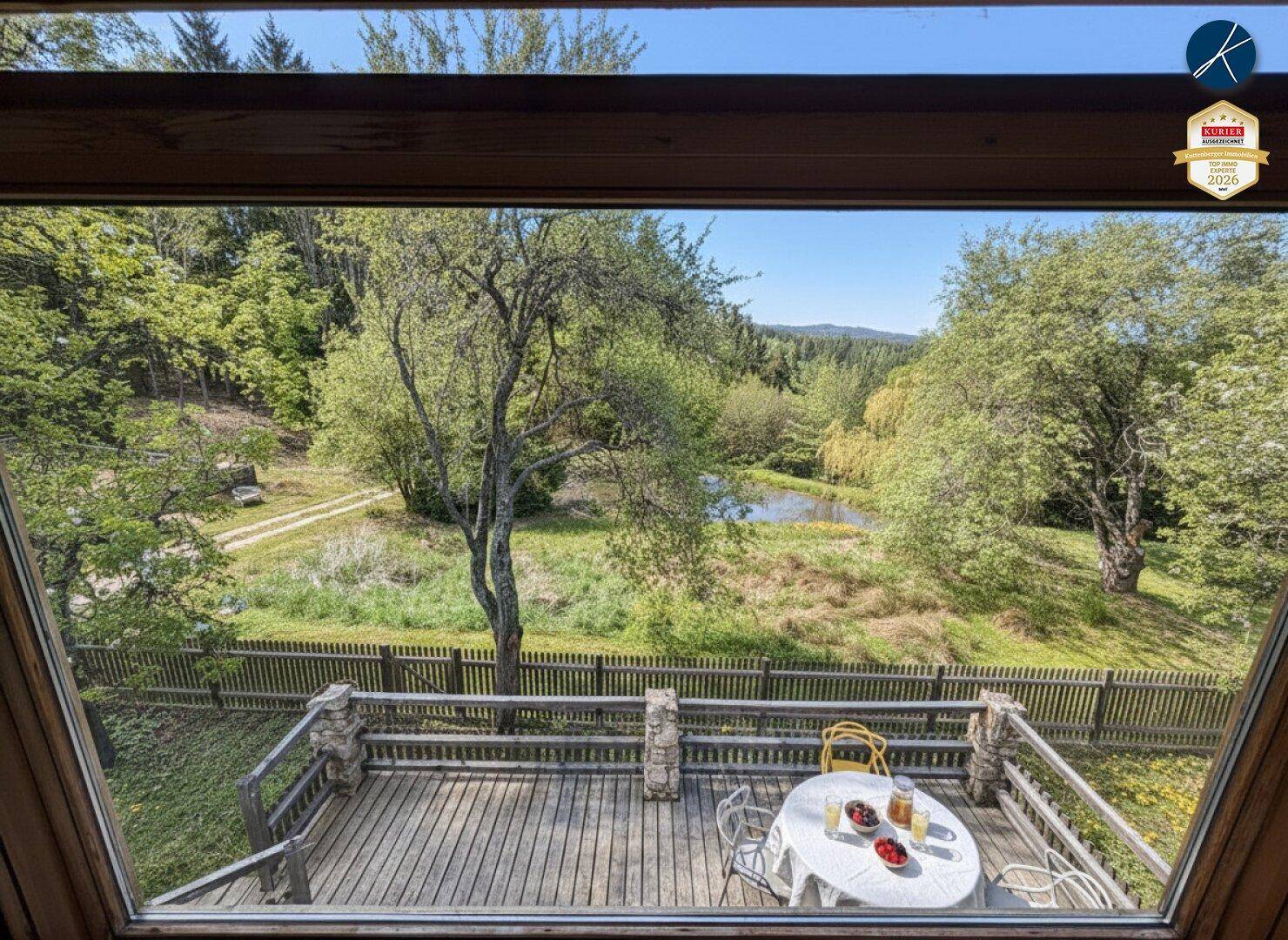 Holzterrasse mit Gartenmöbeln und weitem Blick über die grüne Landschaft und einen Teich.