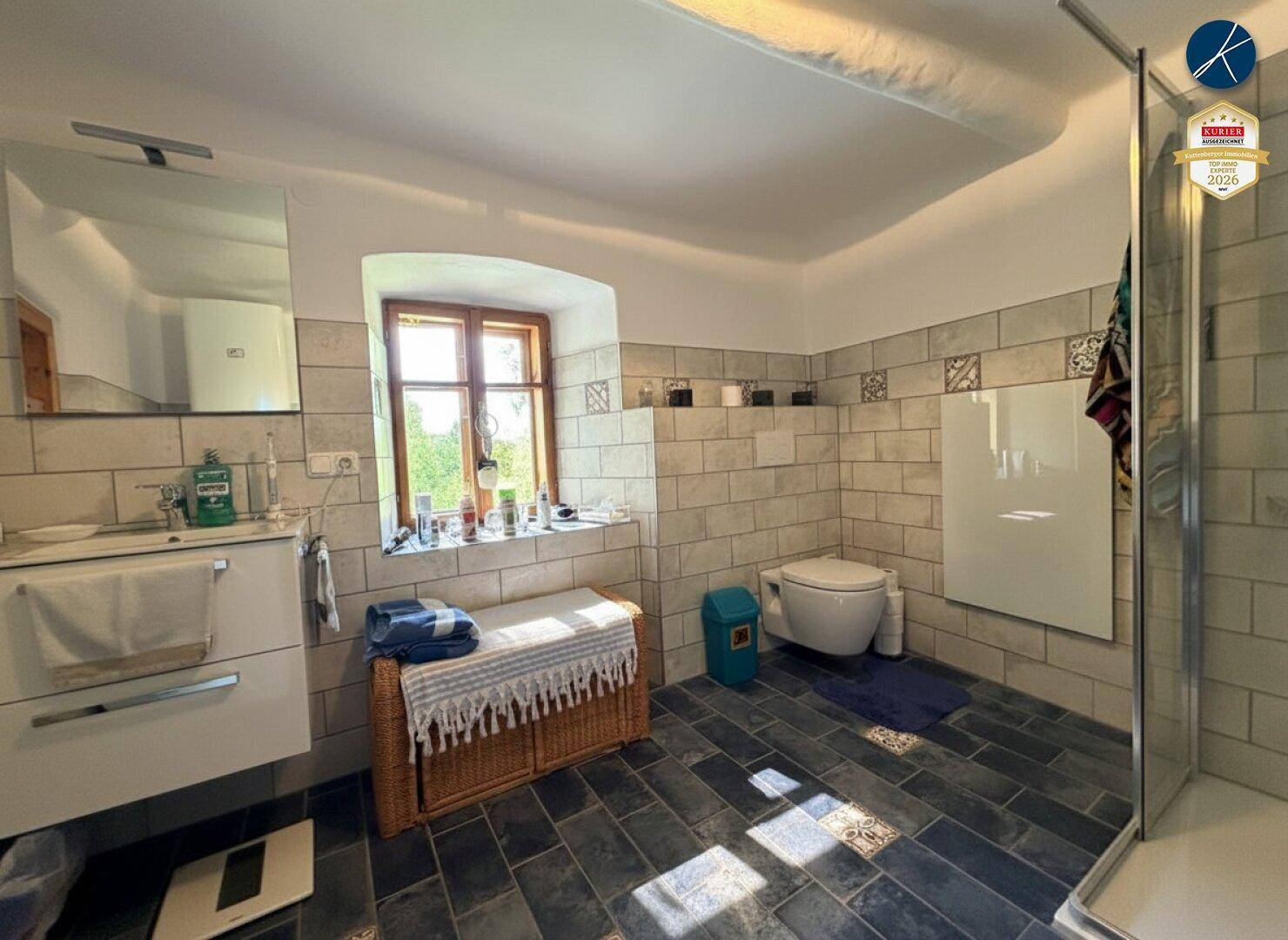 Modernisiertes Badezimmer mit grauen Fliesen, begehbarer Dusche und Fenster.