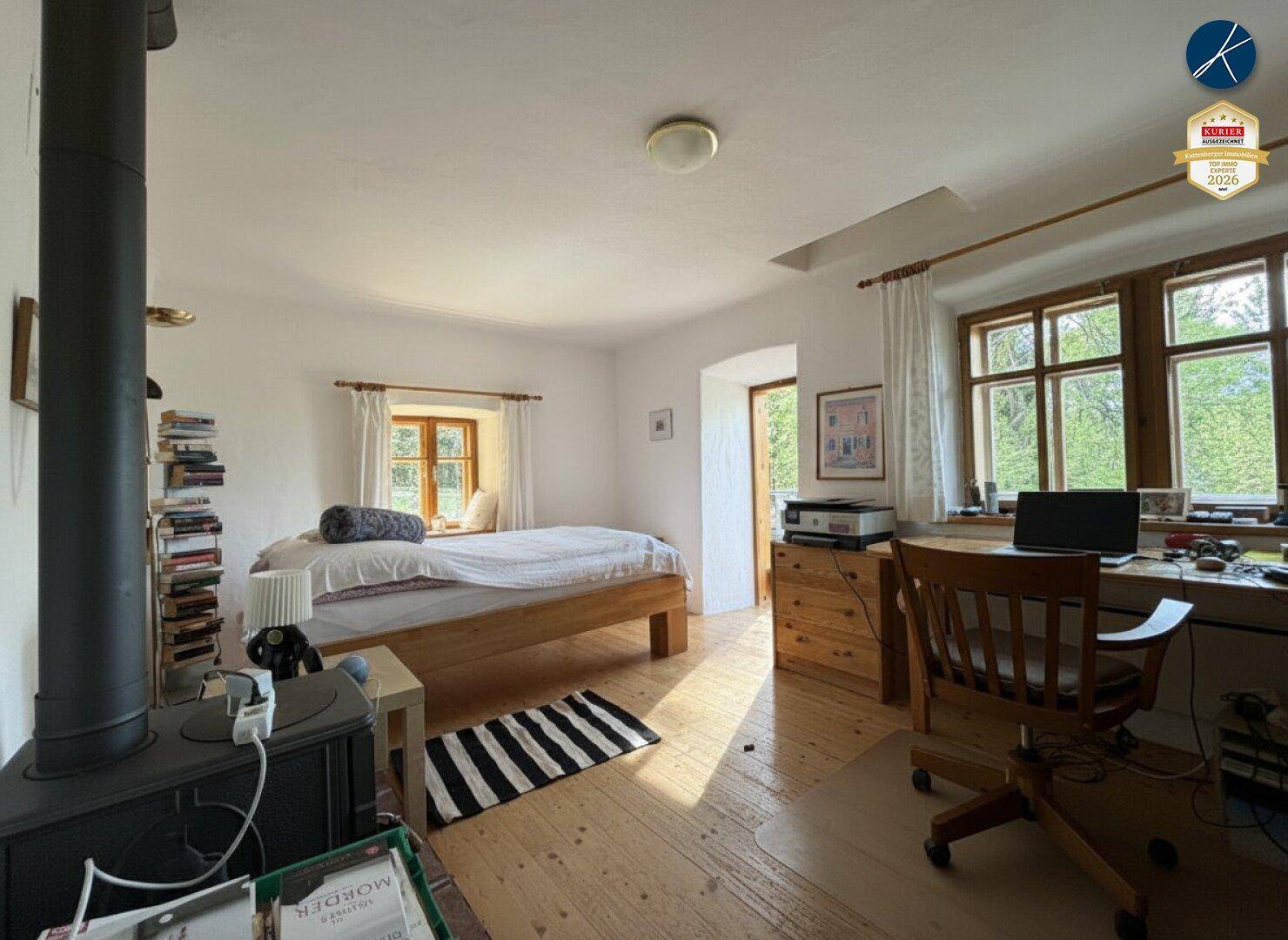 Helles Schlafzimmer mit Holzboden, Kaminofen und Arbeitsplatz am Fenster mit Blick ins Grüne.