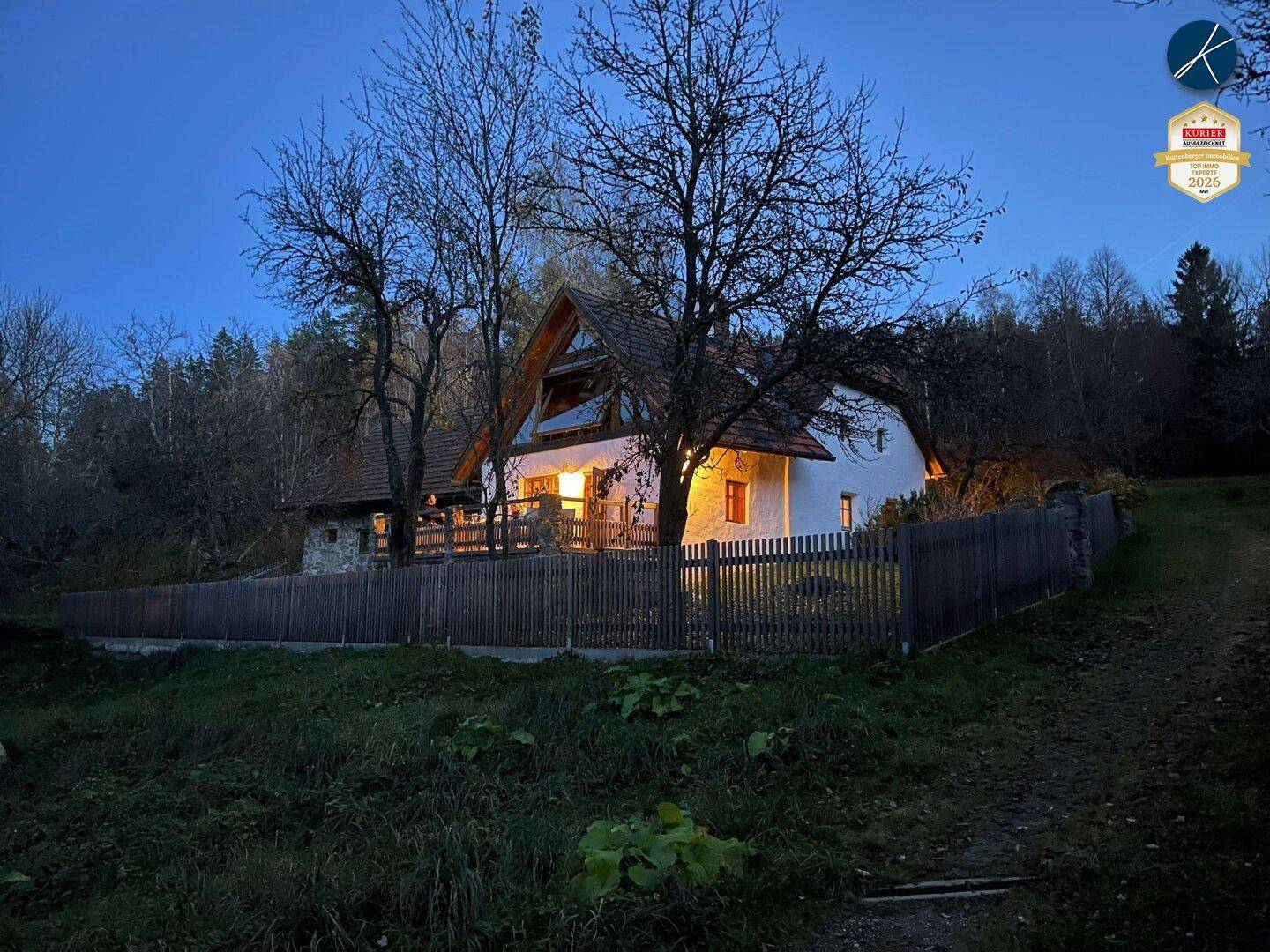 Beleuchtetes Landhaus in der Dämmerung, umgeben von Bäumen und einem Holzzaun.