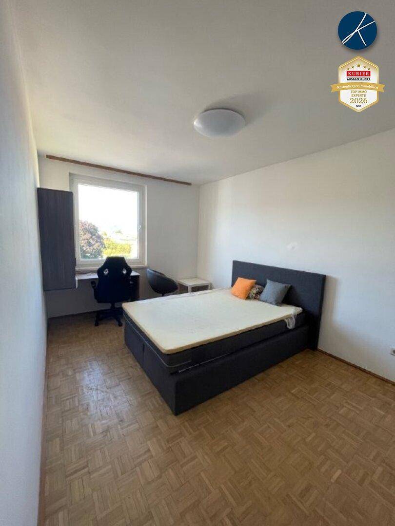 Schlafzimmer mit Holzboden, einem Bett und einem Arbeitsbereich am Fenster, das viel Tageslicht hereinlässt.