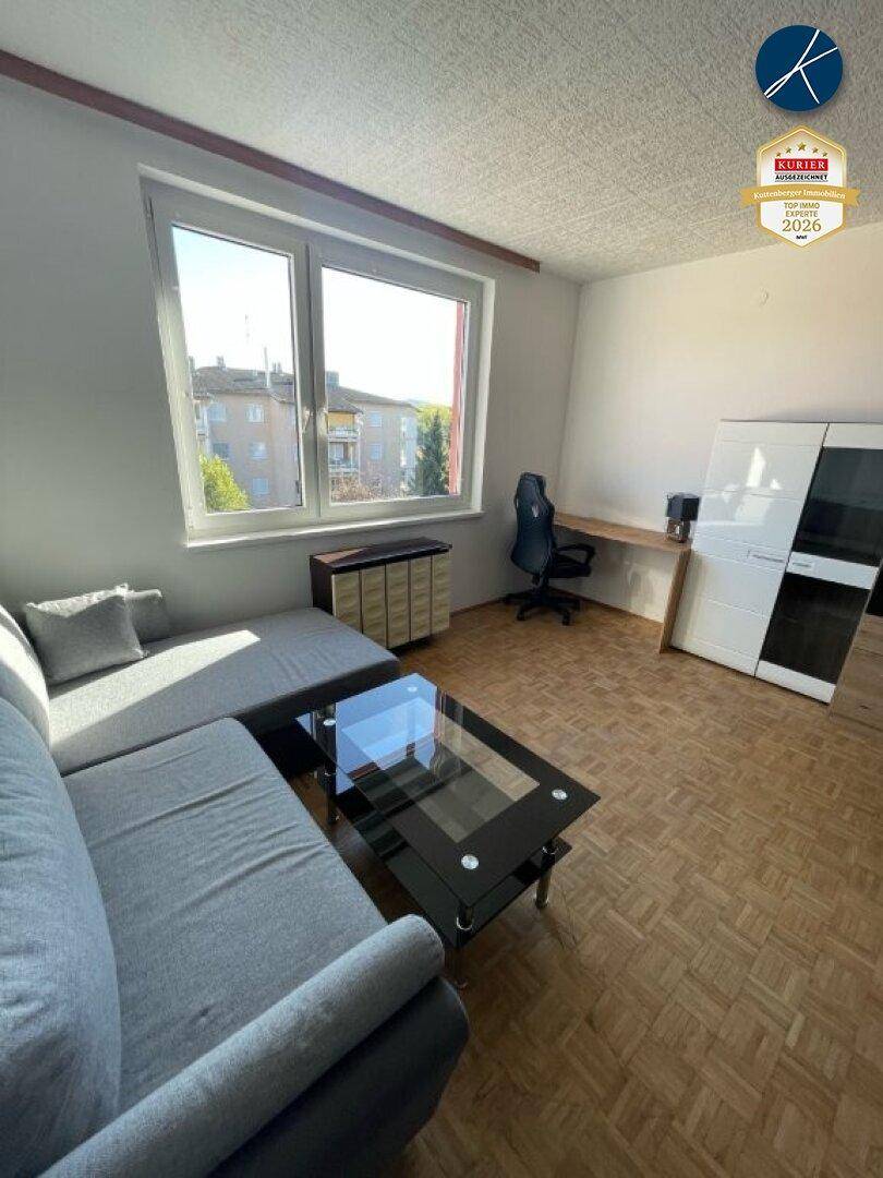 Gemütliches Wohnzimmer mit grauem Sofa, Couchtisch und einem Arbeitsplatz, beleuchtet durch ein großes Fenster.