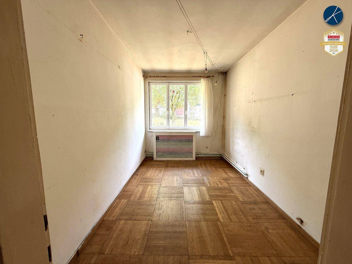 Leeres Zimmer mit Parkettboden und Fenster, das auf den Garten blickt.