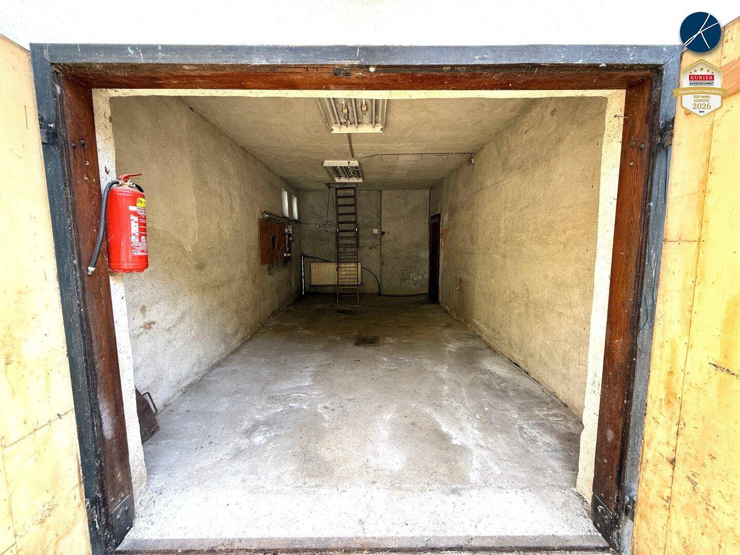 Leere Garage mit Betonboden und Zugang zu einem oberen Bereich über eine Leiter.