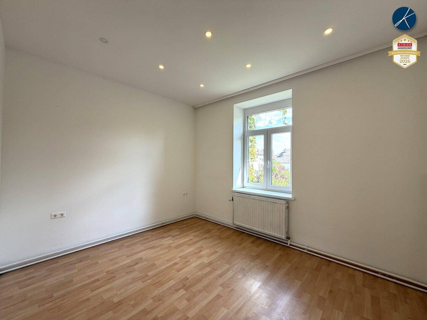 Helles, leeres Schlafzimmer mit Parkettboden, einem Fenster und weiß gestrichenen Wänden.