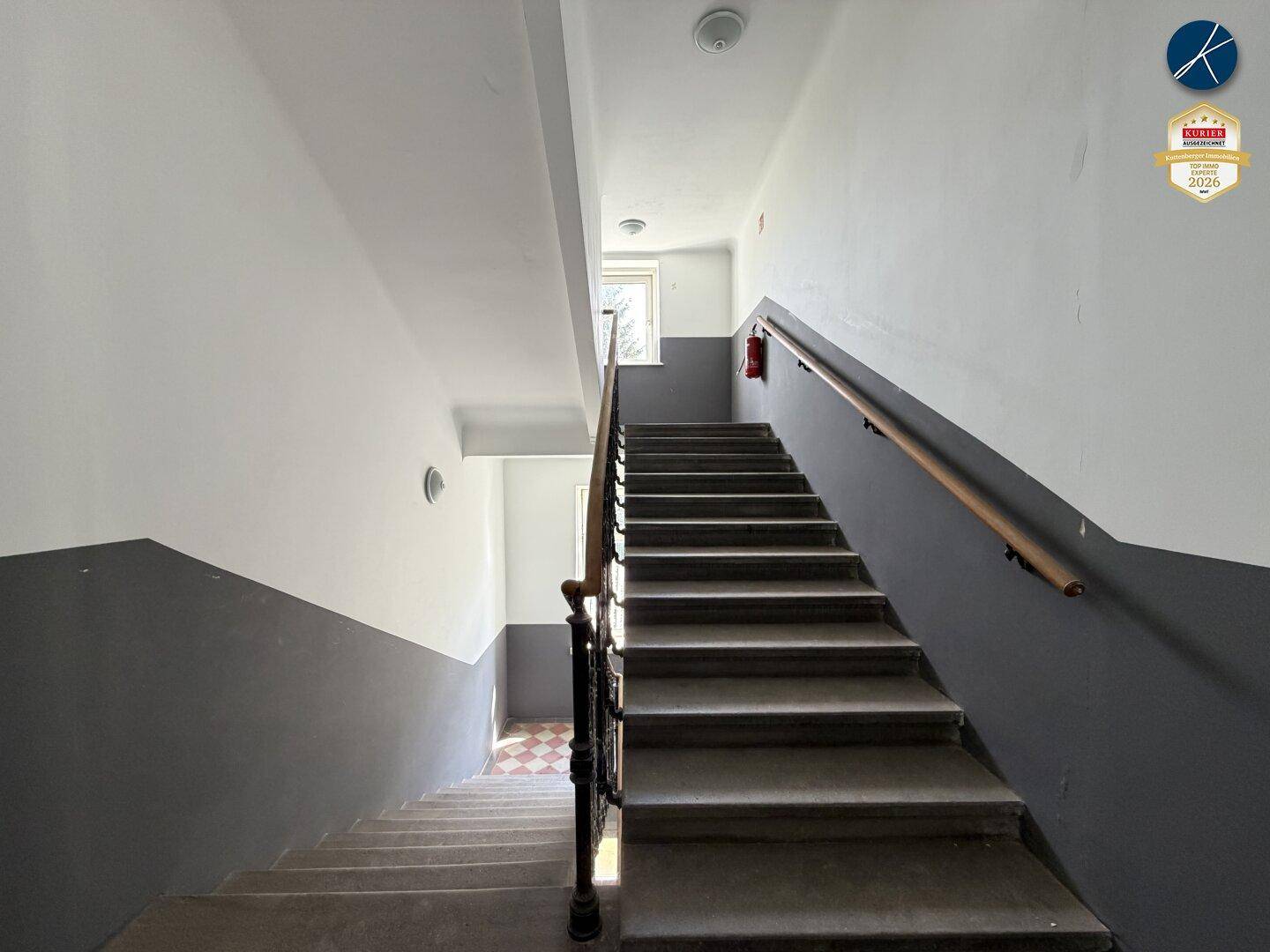 Treppenhaus mit Steinstufen, Holzgeländer und zweifarbigen Wänden in Grau und Weiß.