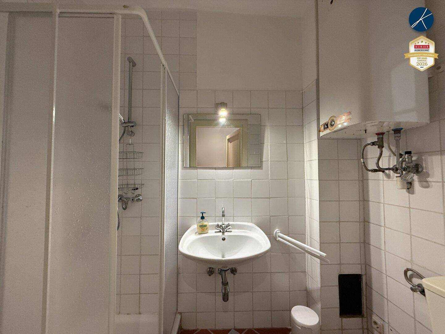 Badezimmer mit weißen Fliesen, Dusche und Waschbecken, ausgestattet mit einem Wandspiegel.