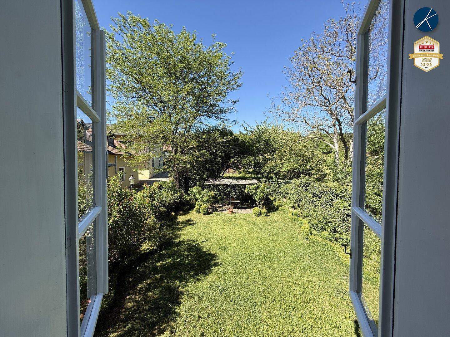 Blick aus dem Fenster in einen grünen, gepflegten Garten mit Rasenfläche und Bäumen unter blauem Himmel.