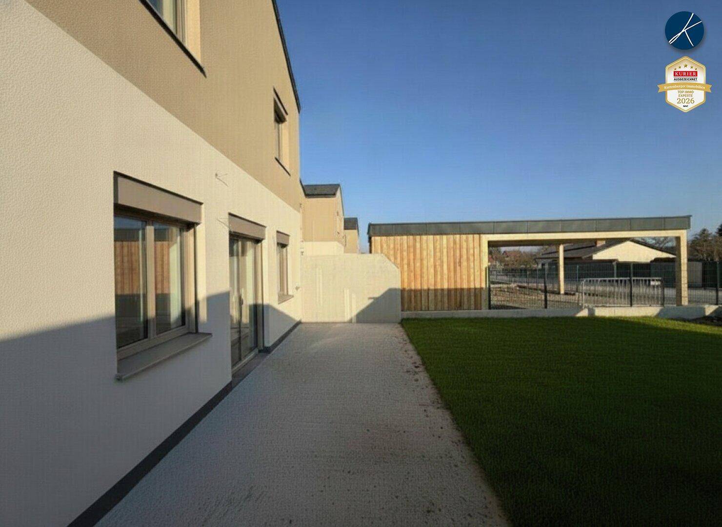 Seitenansicht eines Reihenhauses mit gepflastertem Weg, Rasenfläche und Carport im Hintergrund.