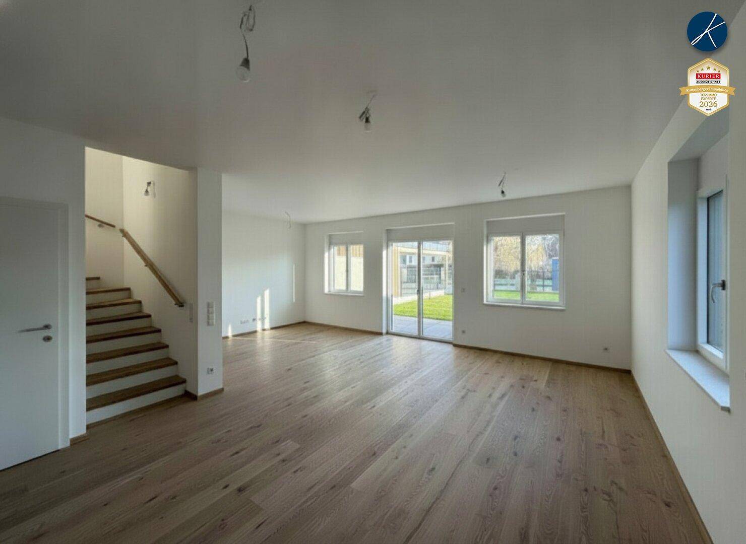 Helles, leeres Wohnzimmer mit Holztreppe, großen Fenstern und Parkettboden.