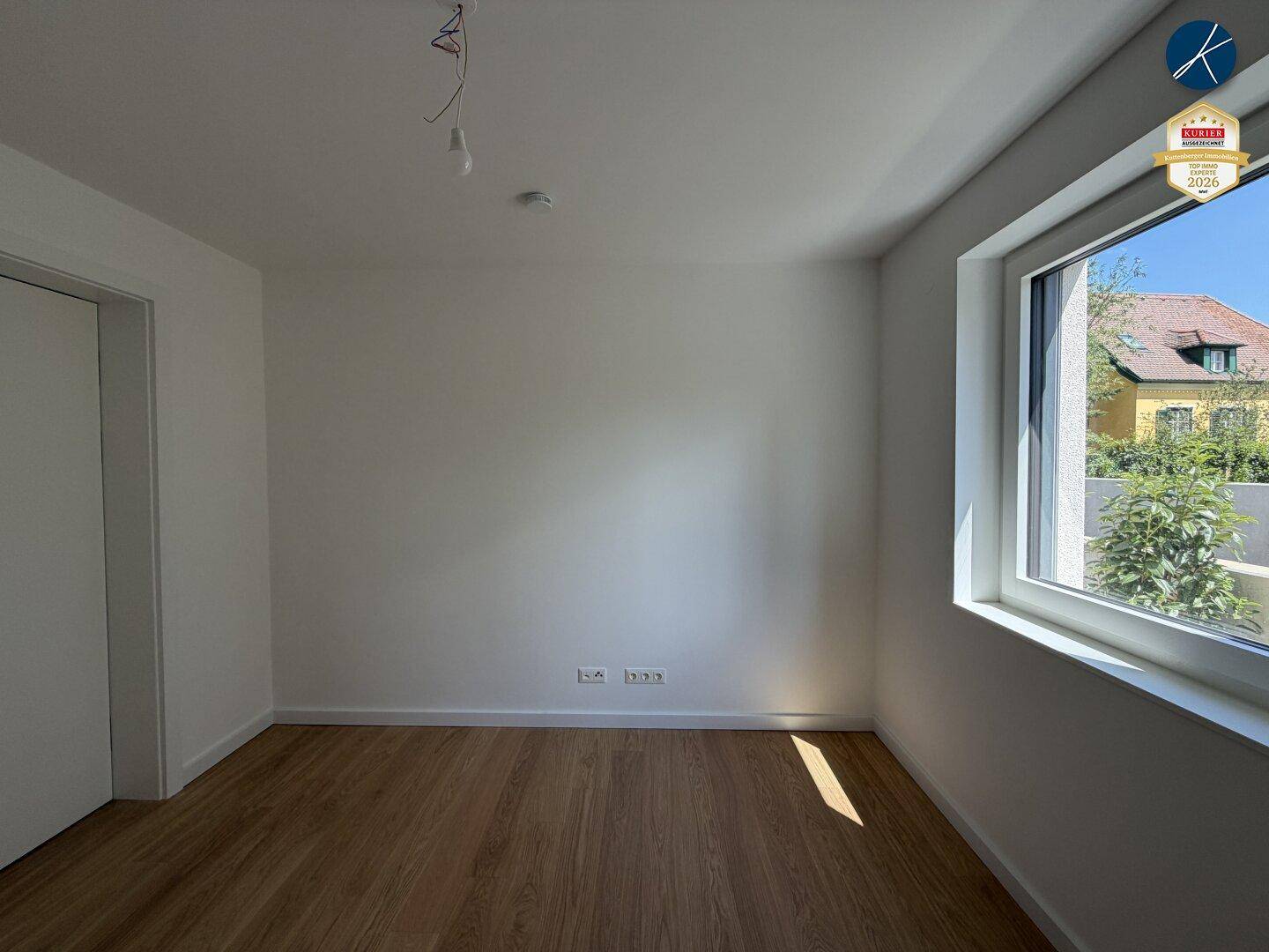 Helles, leeres Zimmer mit Holzboden und Fenster mit Blick ins Grüne.