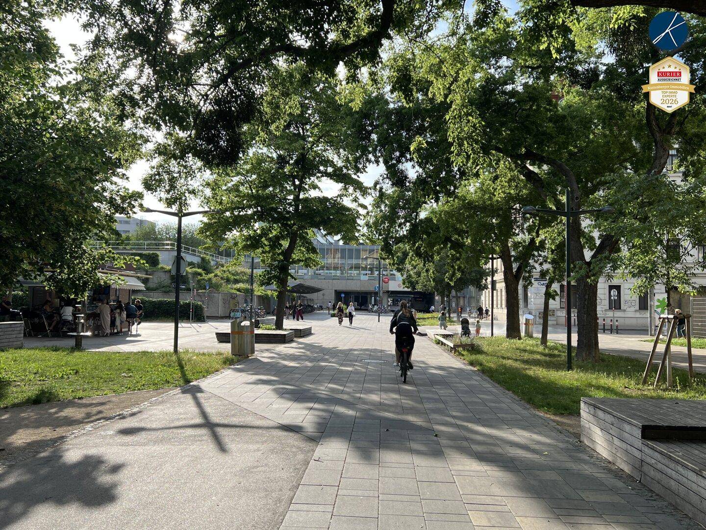 Grüner Fußgängerweg mit Bäumen und Radfahrern in einer städtischen Umgebung.