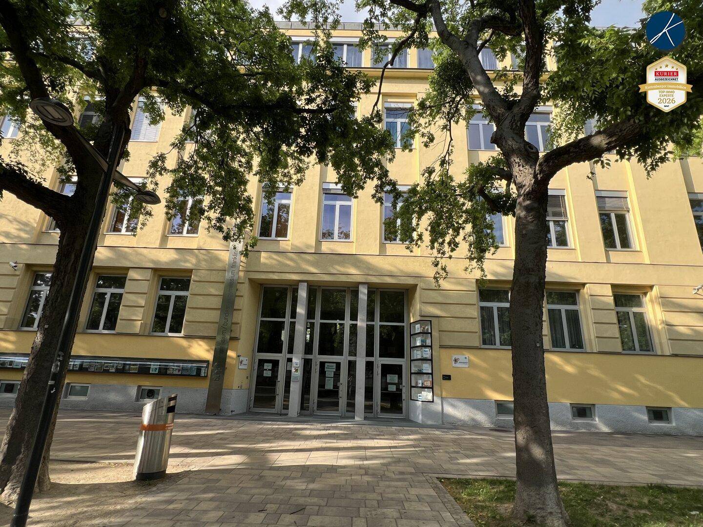 Gelbes Gebäude mit vielen Fenstern und einem großen Glaseingang, umgeben von Bäumen.