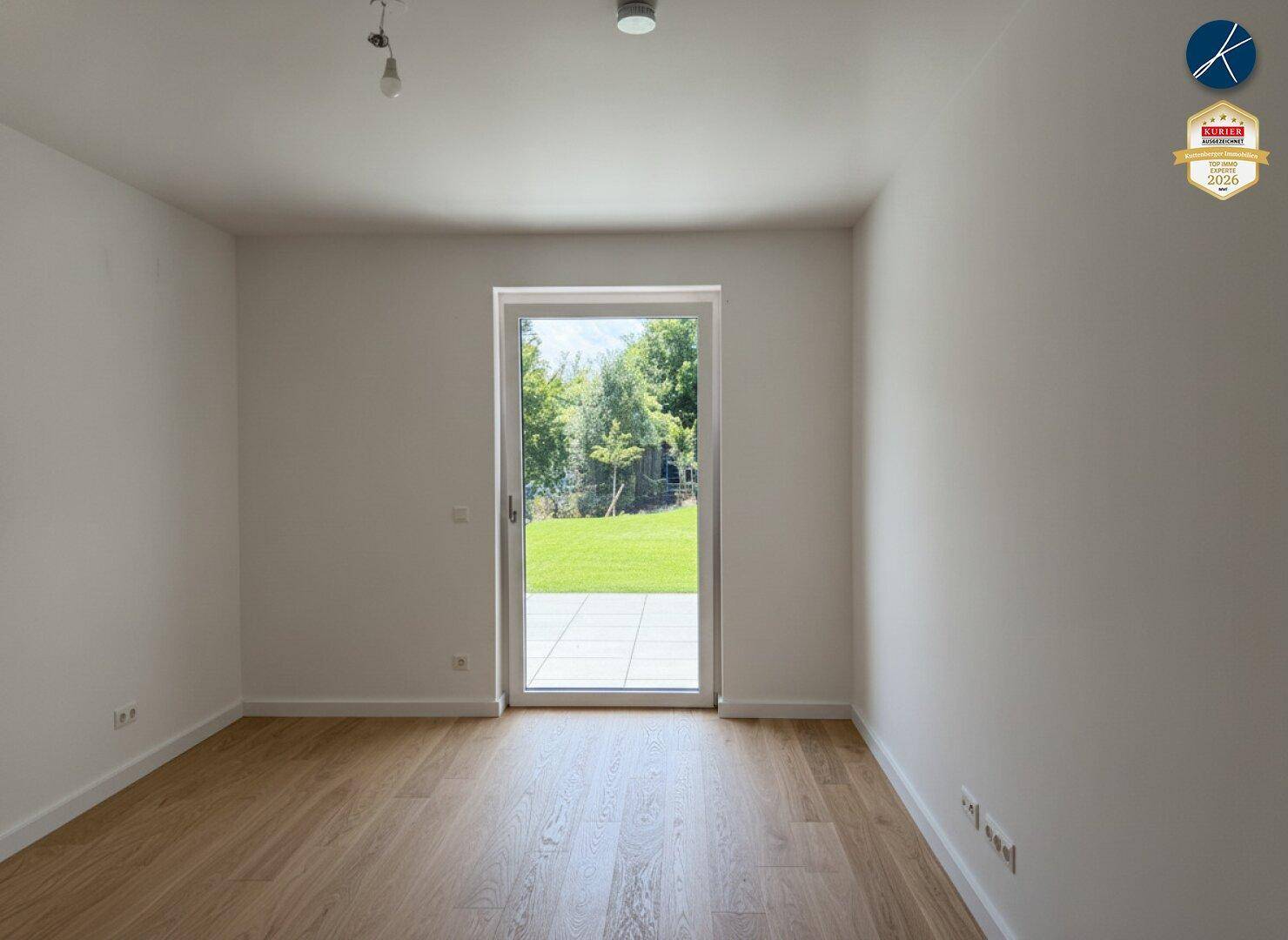Helles, leeres Zimmer mit Holzboden und direktem Zugang zum Garten durch eine Glastür.