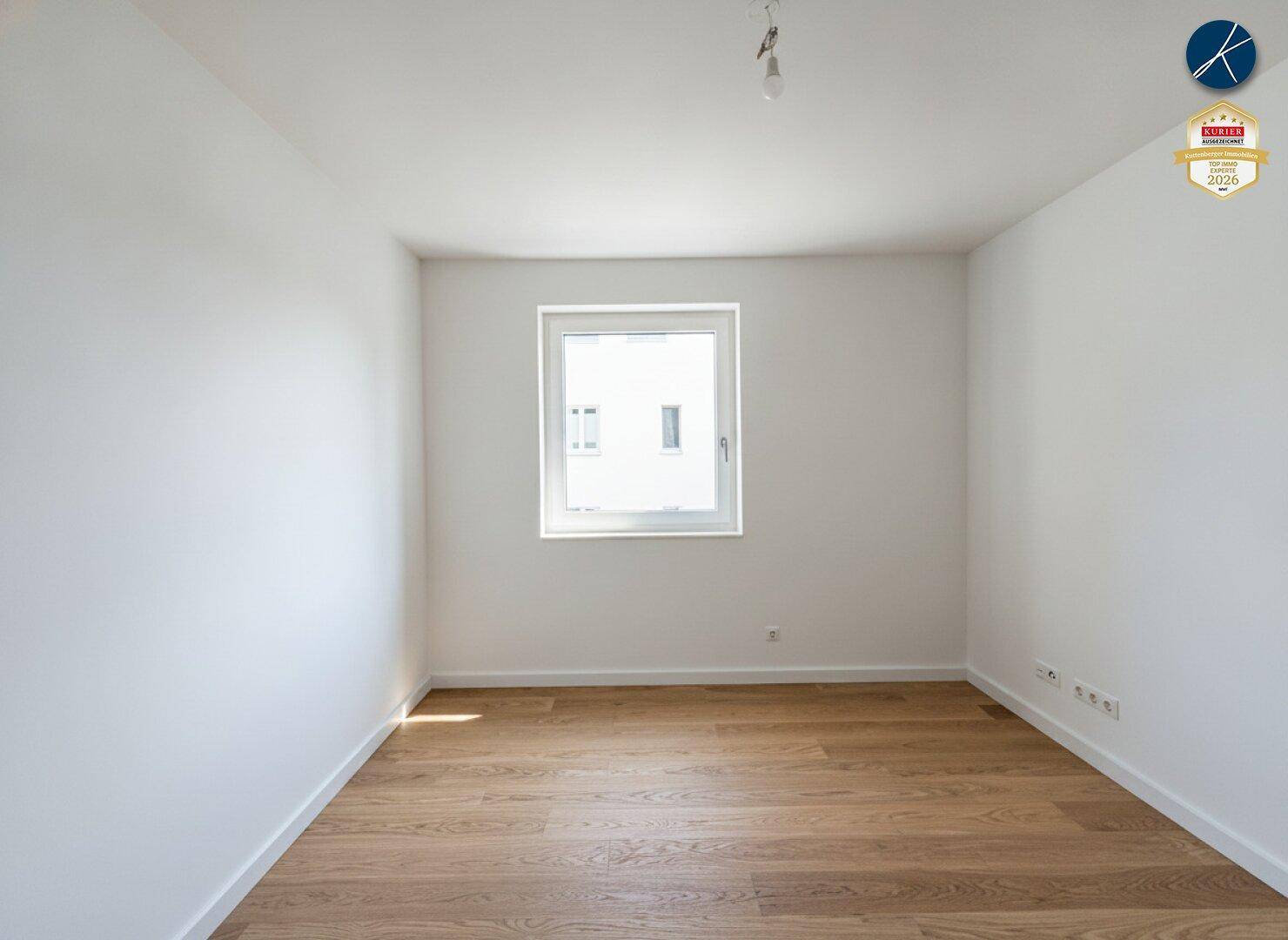 Schlichtes Schlafzimmer mit Parkettboden und einem Fenster, das viel Tageslicht hereinlässt.