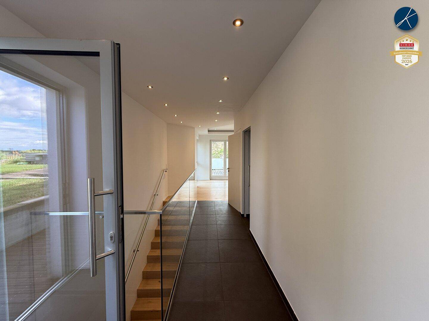 Heller Flur mit moderner Glasbrüstung an der Treppe, Einbaustrahlern und dunklem Fliesenboden.