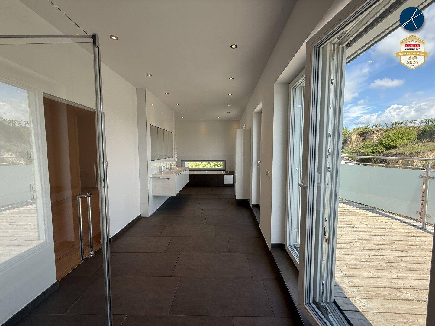 Langes Badezimmer mit dunklem Fliesenboden, Doppelwaschbecken und Zugang zu einer Holzterrasse.