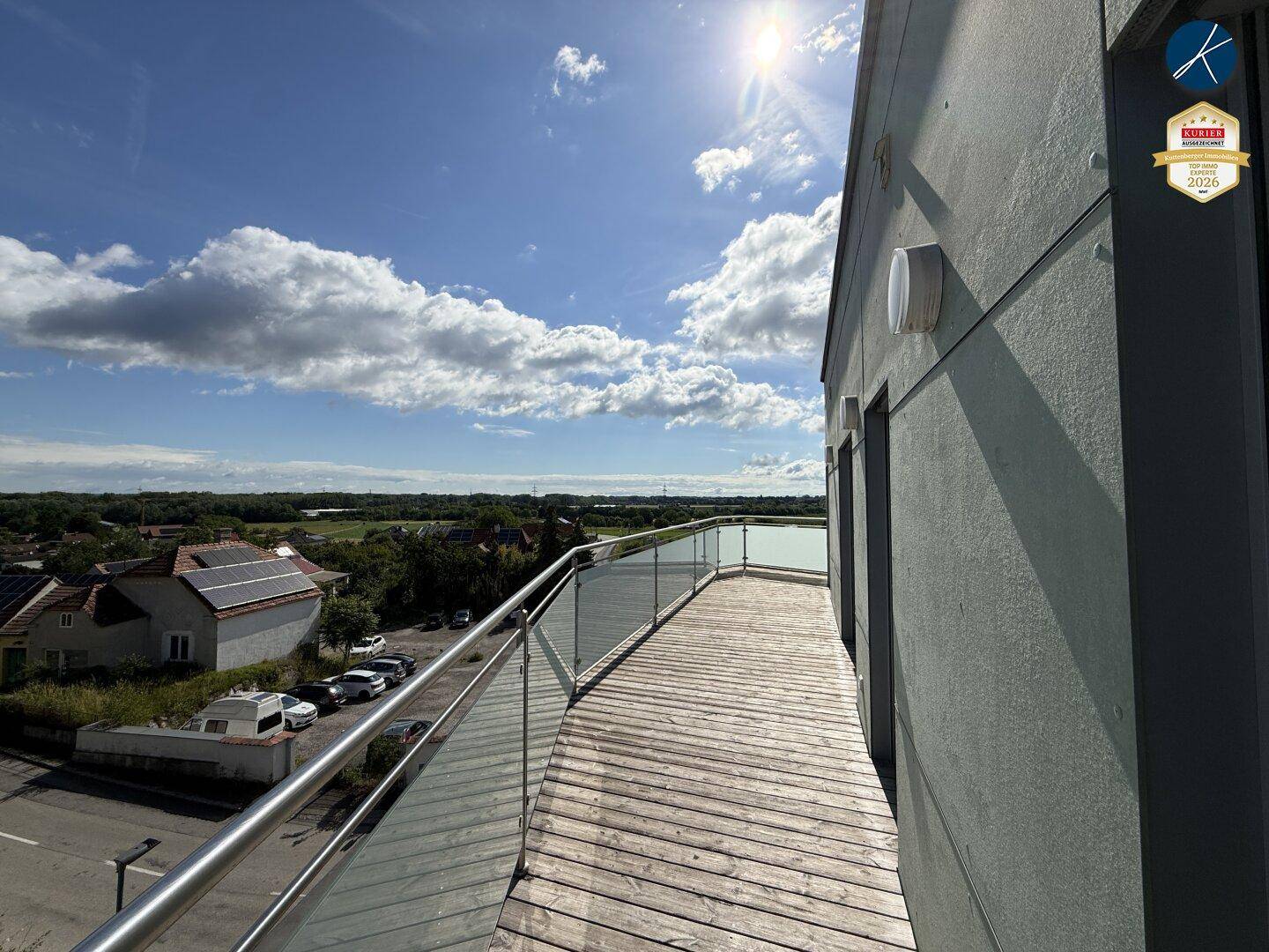 Langer Holzbalkon mit Glasgeländer und weitem Blick über die ländliche Umgebung unter blauem Himmel.