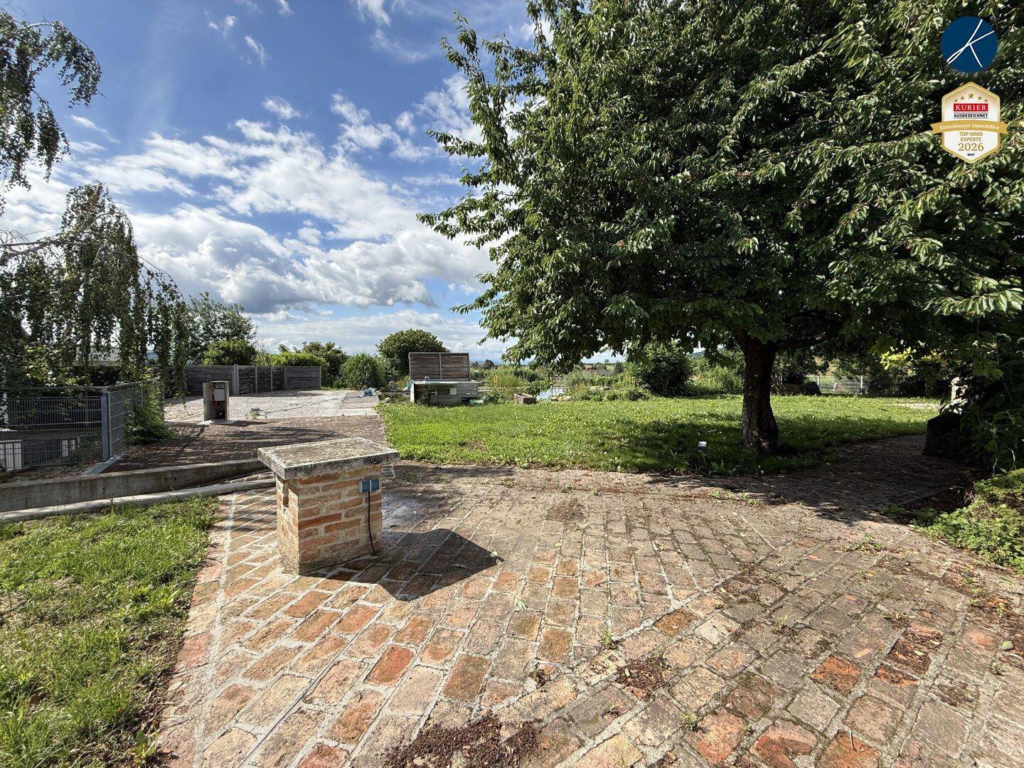 Sonniger Gartenbereich mit Ziegelpflaster, Rasenfläche und altem Baumbestand unter blauem Himmel.