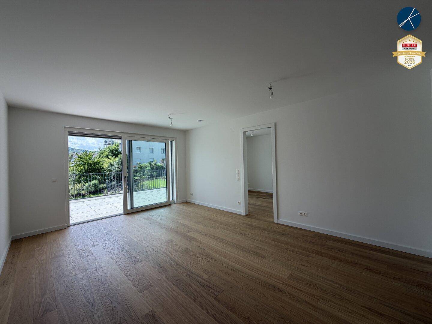 Heller, leerer Wohnraum mit Holzboden und Zugang zu einer großen Terrasse.