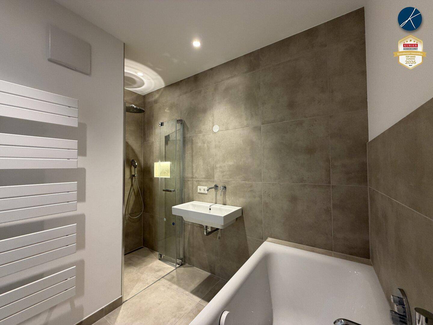 Modernes Badezimmer mit Dusche, Badewanne, beheiztem Handtuchhalter und grauen Fliesen.